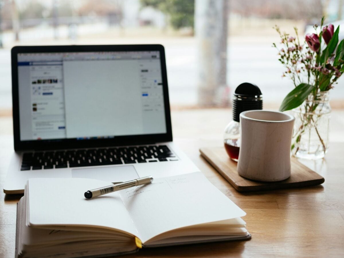 tablet with keyboard sitting on a desk with a cup of coffee, flower arrangement, and an opened book with a writing pen on top.
